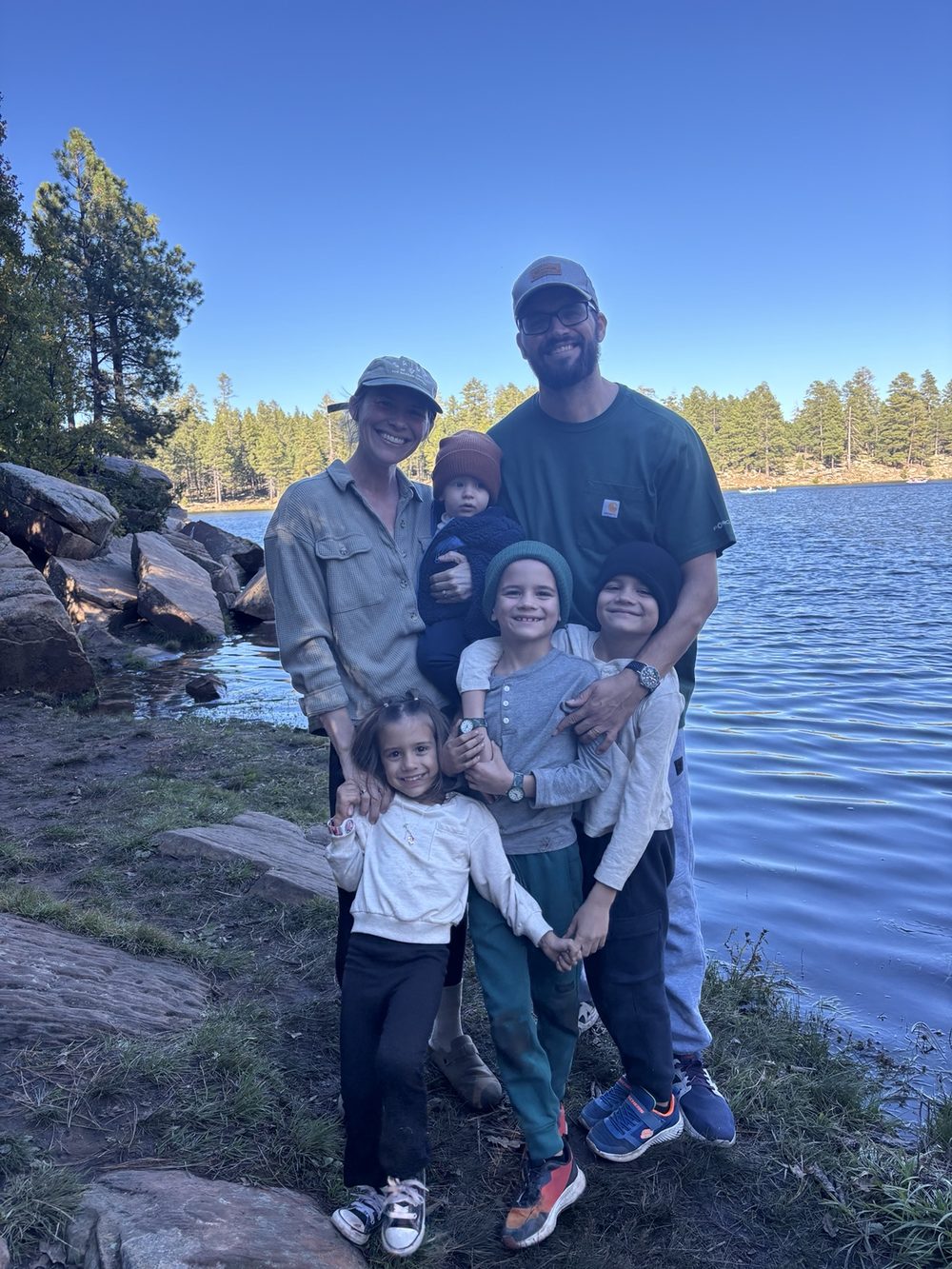 Our family of seven smiling together on the shore of a pine-lined lake